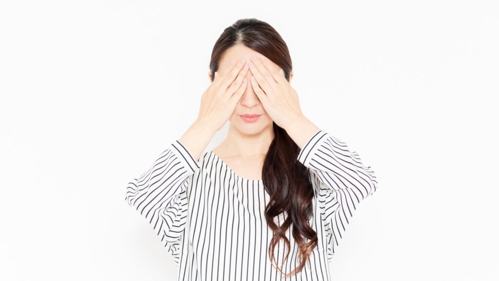 白い背景の前で、ストライプ柄の服を着て両手で自分の目を覆い隠す女性
