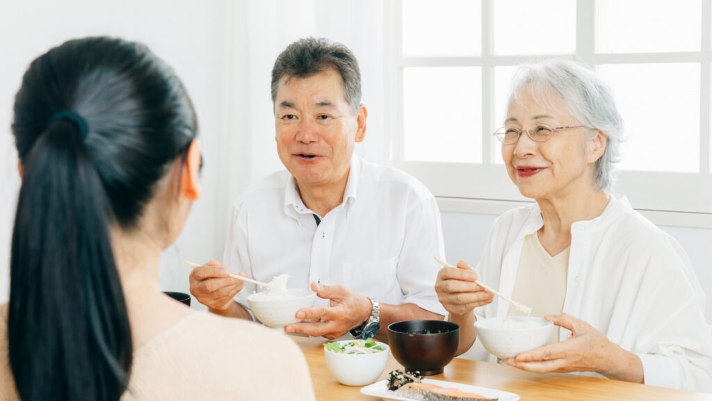 食卓でご飯を食べながら、手前の女性と笑顔で会話をするシニア世代の夫婦