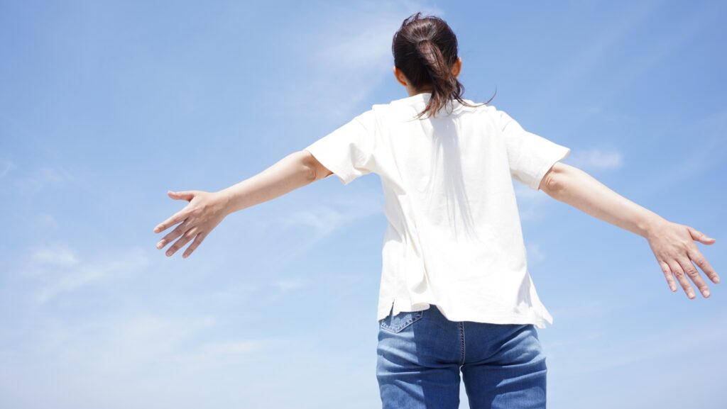 青空の下で両手を大きく広げて爽やかな解放感を感じている女性の後ろ姿
