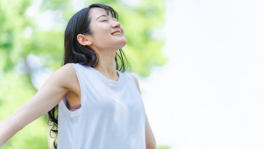 新緑の木々を背景に、目を閉じて笑顔で深呼吸をする白いノースリーブ姿の女性