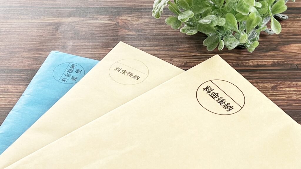 木目調のテーブルの上に重ねられた、「料金後納」のスタンプが押された複数の茶封筒と青い封筒