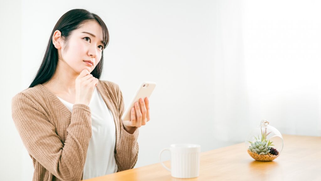 室内のテーブルでマグカップの横に座り、スマートフォンを片手に持ちながら悩ましげに考え事をする女性