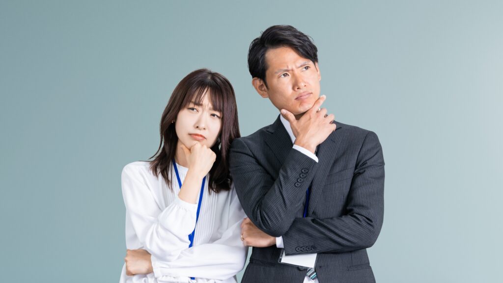 グレーの背景の前で、顎に手を当てて困った表情で斜め上を見つめる、スーツ姿の男性と社員証を下げた女性