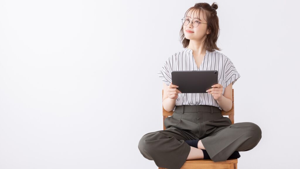 白い背景のなか、木製の椅子の上であぐらをかいて座り、タブレット端末を持ちながら笑顔で斜め上を見つめる眼鏡をかけた女性