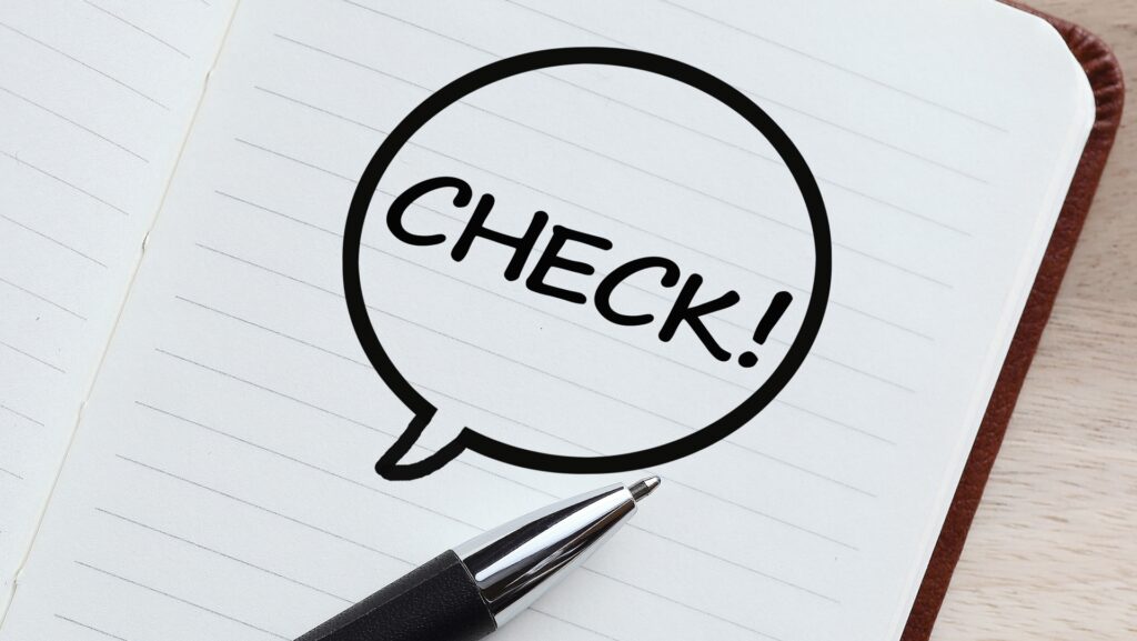 開いたノートの紙面に描かれた「CHECK!」の文字が入った吹き出しと、手前に置かれたボールペン